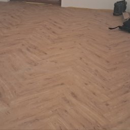 May bud - Ułożona podłoga z paneli w jodełkę, widok z góry. Widoczne narzędzia i akcesoria montażowe. Jasne ściany, gniazdko elektryczne.