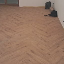 Mat bud - Ukończone układanie paneli podłogowych w jodełkę, widoczne narzędzia pracy przy ścianie, świeżo wykończone wnętrze.