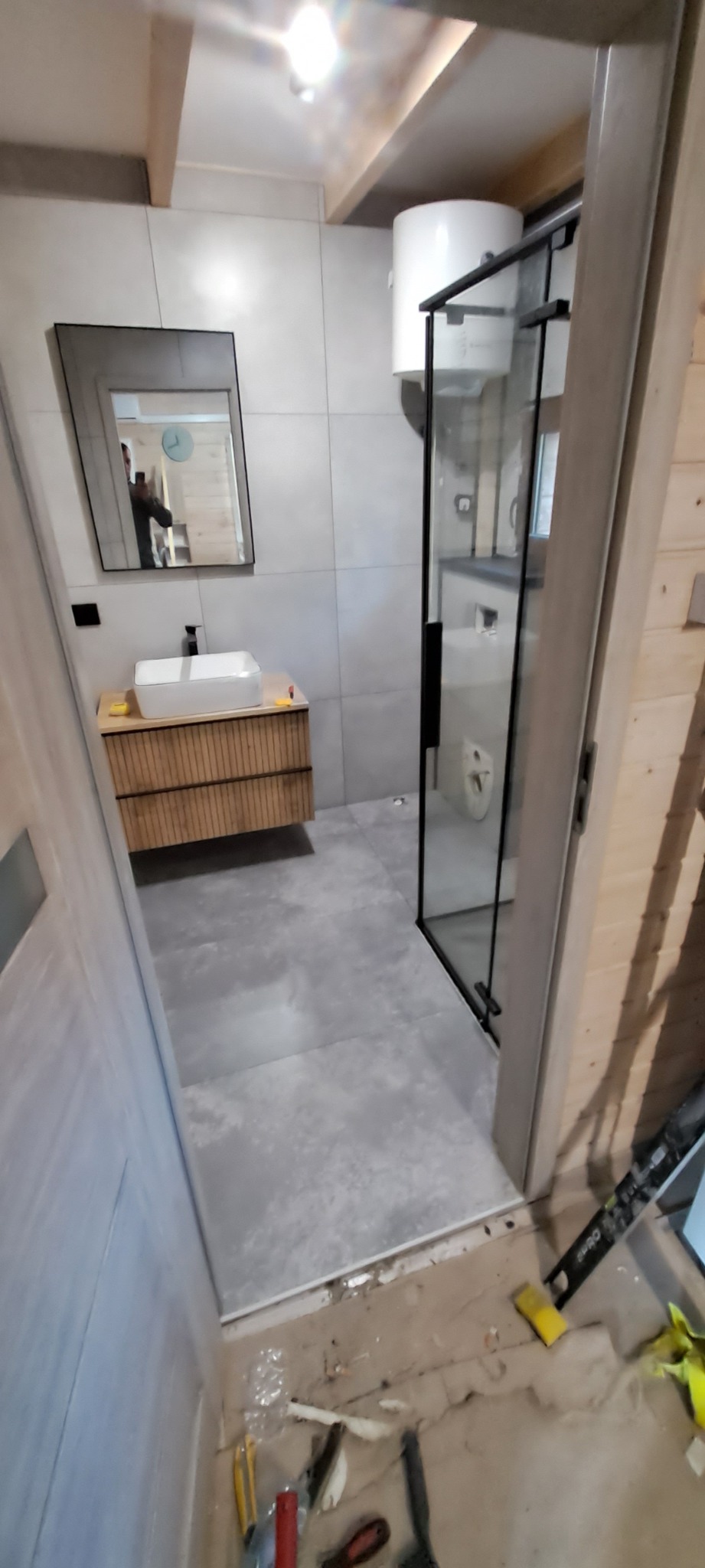 Wykończona łazienka w stylu skandynawskim: szare płytki, drewniana szafka pod umywalką, kabina prysznicowa z czarnymi ramami. Widoczne narzędzia na podłodze.