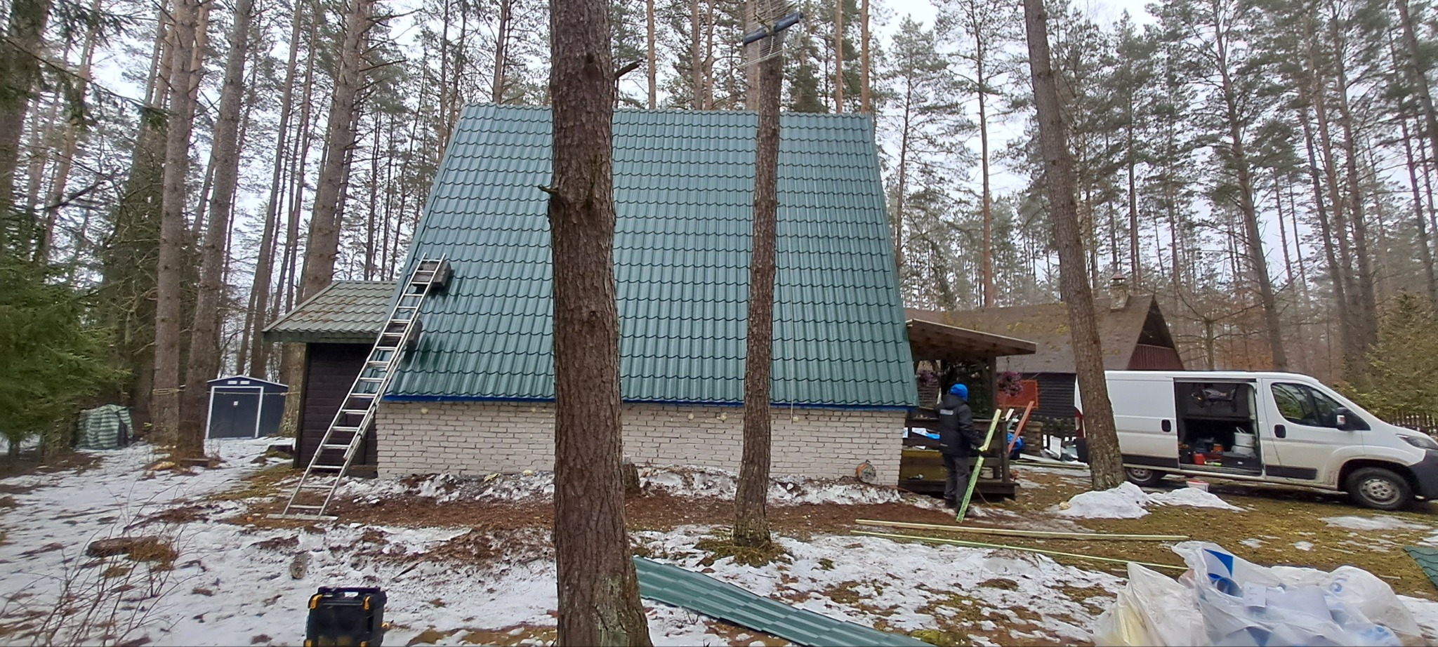 Dom z nowym, zielonym dachem z blachodachówki, widoczna drabina i pracownik. Prace dekarskie w zimowej scenerii, na tle lasu iglastego i zaparkowanego busa.