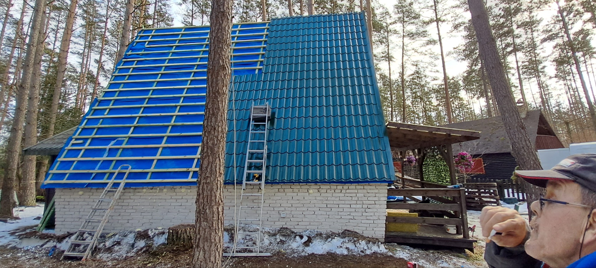 Dach domu w trakcie krycia: połowa z niebieską dachówką, druga z folią i łatami. Drabina oparta o ścianę. W tle las i fragment osoby.