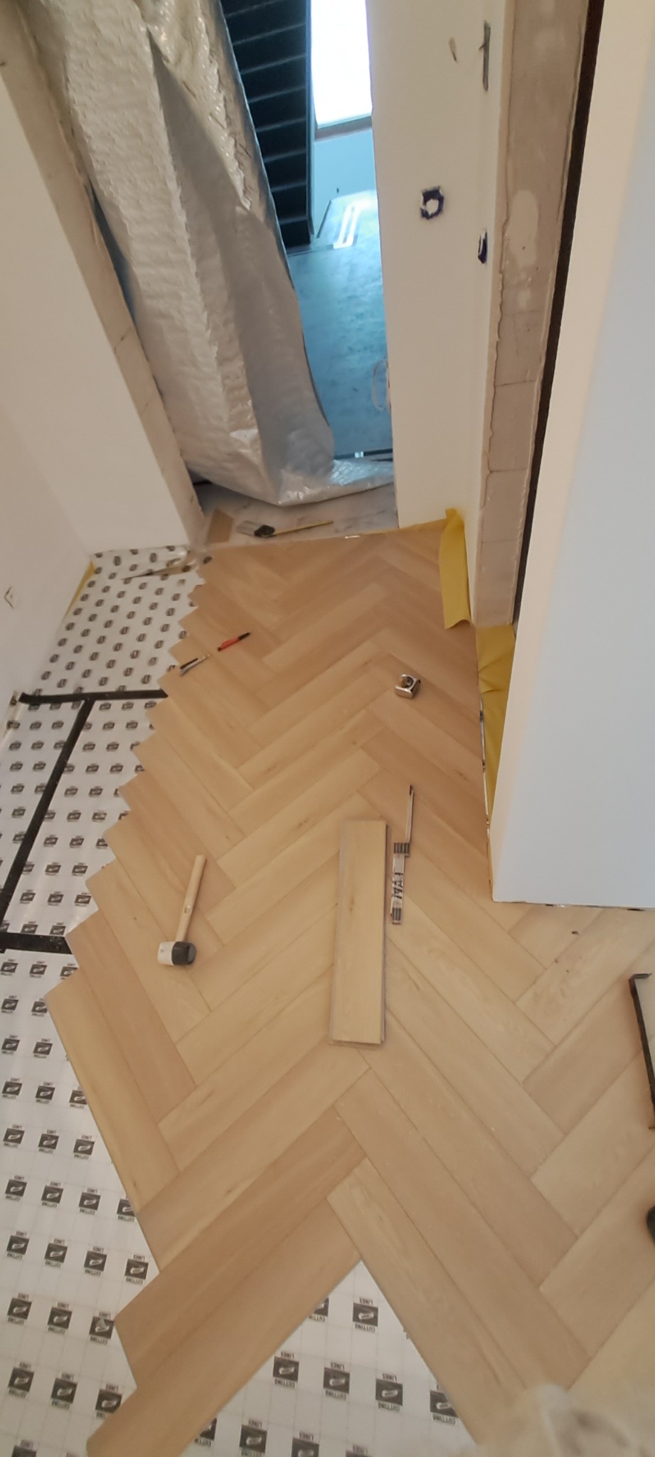 Układanie parkietu w jodełkę na podkładzie. Widoczne narzędzia: młotek, miarka, ołówek. Częściowo ułożona podłoga z jasnego drewna. Widok z góry.