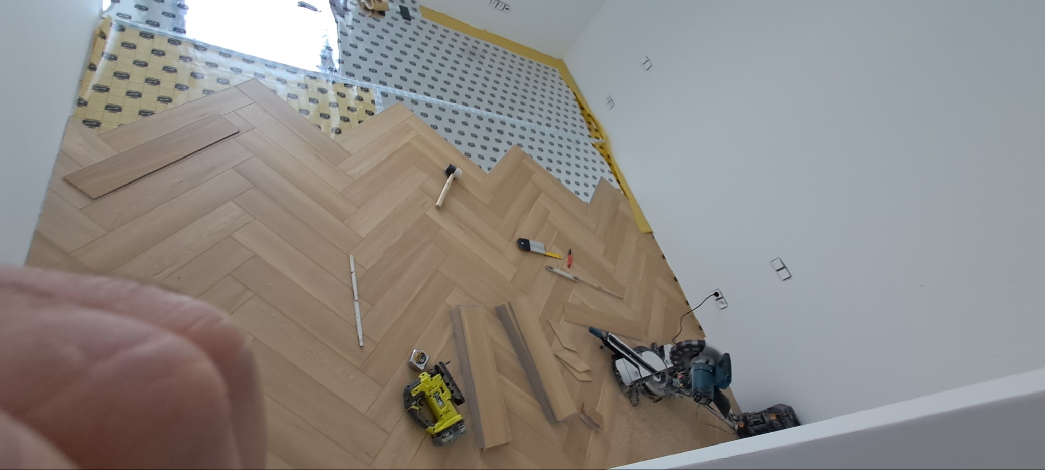 Układanie parkietu w jodełkę: widok z góry na częściowo ułożoną podłogę, narzędzia pracy (młotek, miarka, piła) i podkład pod panele. Ujęcie z bliska, z ręką w kadrze.