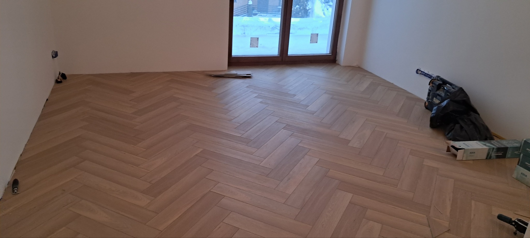 Ułożony parkiet w jodełkę francuską w jasnym odcieniu drewna, widoczne narzędzia i resztki materiałów po montażu, okno z widokiem na zimowy krajobraz.