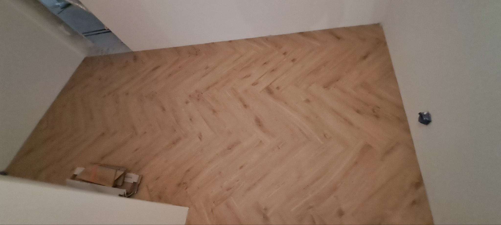 Ściana wyłożona panelami w jodełkę, widoczne kartony po panelach przy podłodze. Białe ściany i sufit. Ujęcie z dołu, lekko z ukosa.