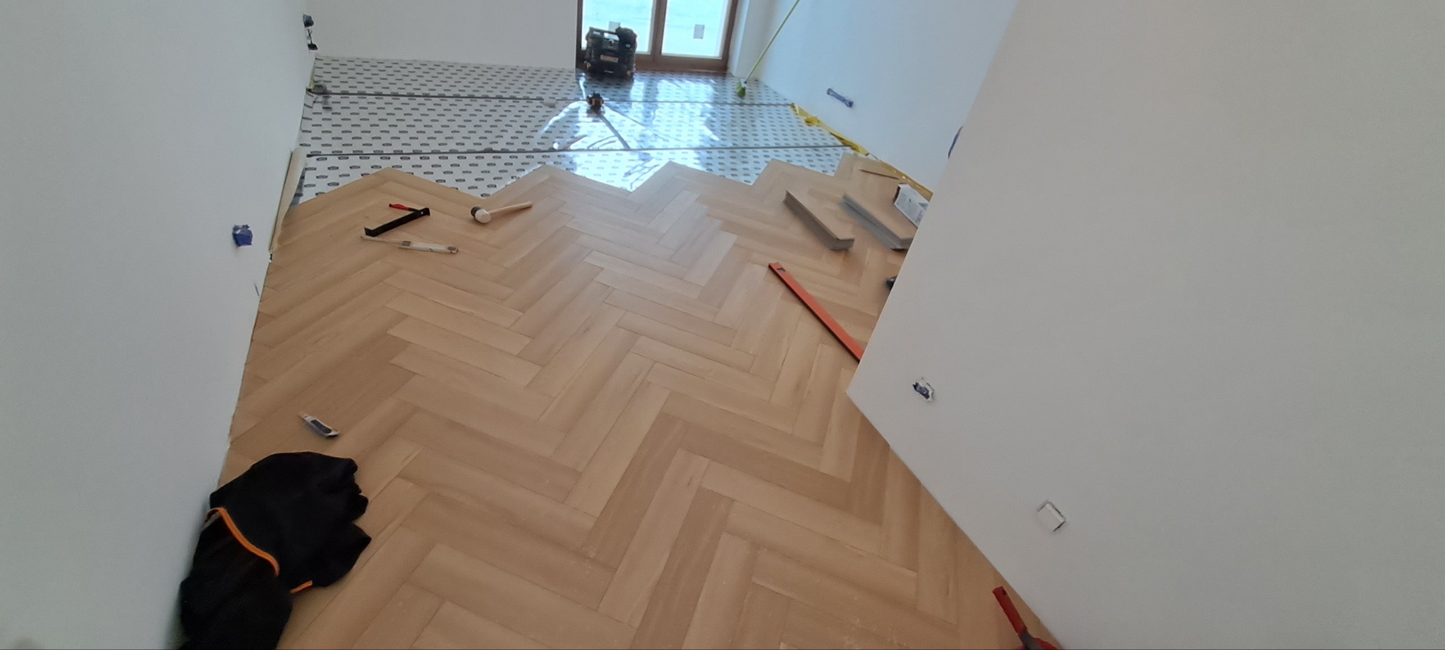 Układanie parkietu w jodełkę: widok z góry na częściowo ułożoną podłogę, narzędzia pracy i podkład. W tle okno i sprzęt budowlany. Jasne wnętrze w trakcie prac wykończeniowych.