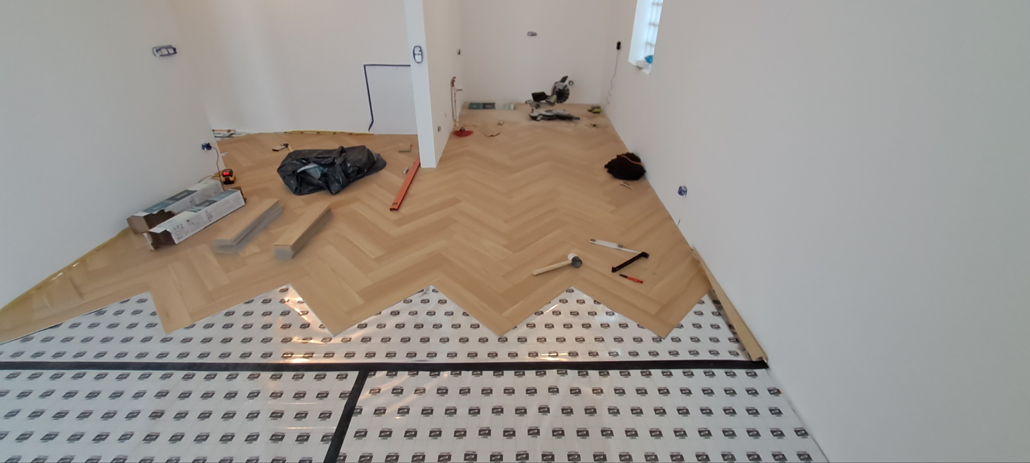 Układanie parkietu w jodełkę na podkładzie izolacyjnym. Widoczne narzędzia, resztki materiałów i częściowo ułożona podłoga w jasnym, nowym pomieszczeniu.