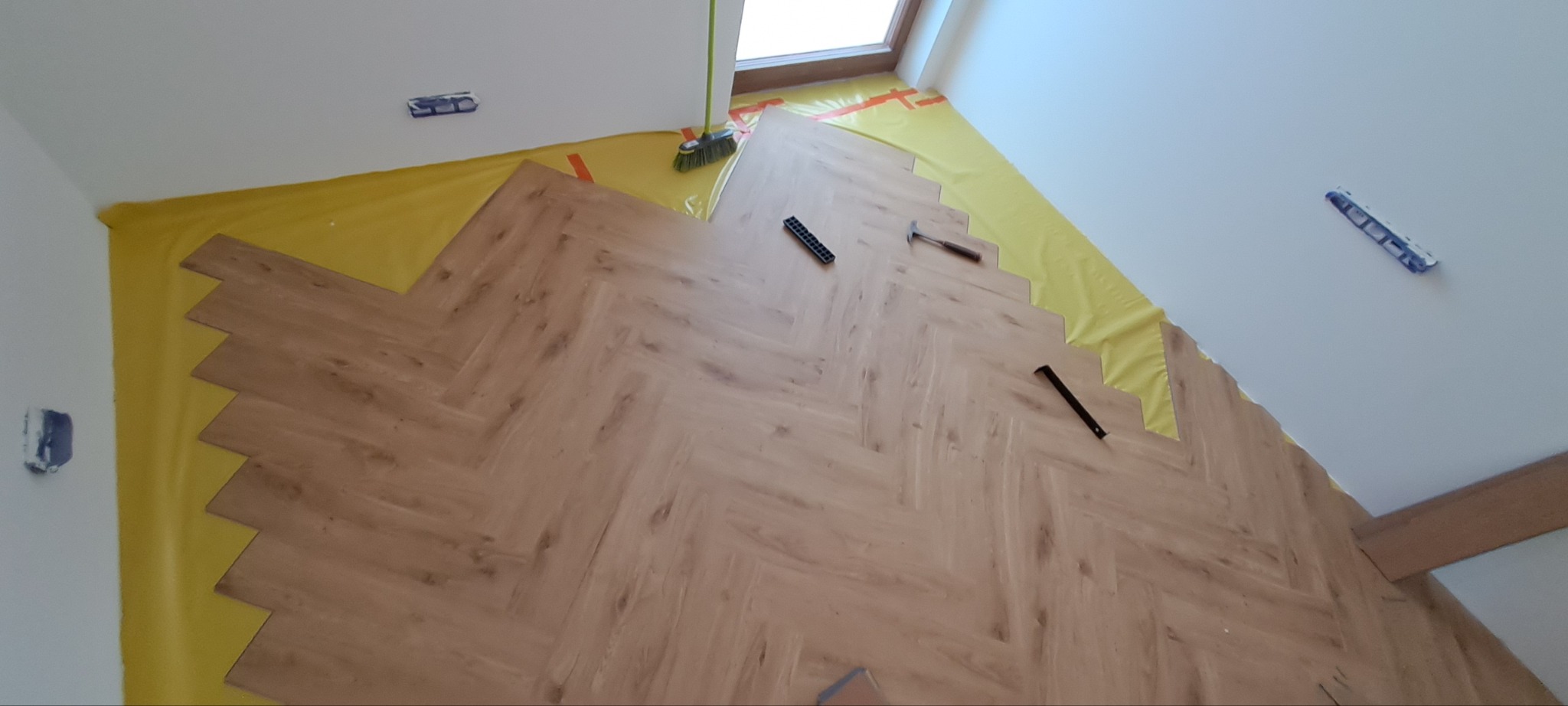 Układanie parkietu w jodełkę na żółtej folii. Widoczne narzędzia: młotek, listwa, podkładka. Częściowo ułożona podłoga w jasnym pomieszczeniu z oknem dachowym.