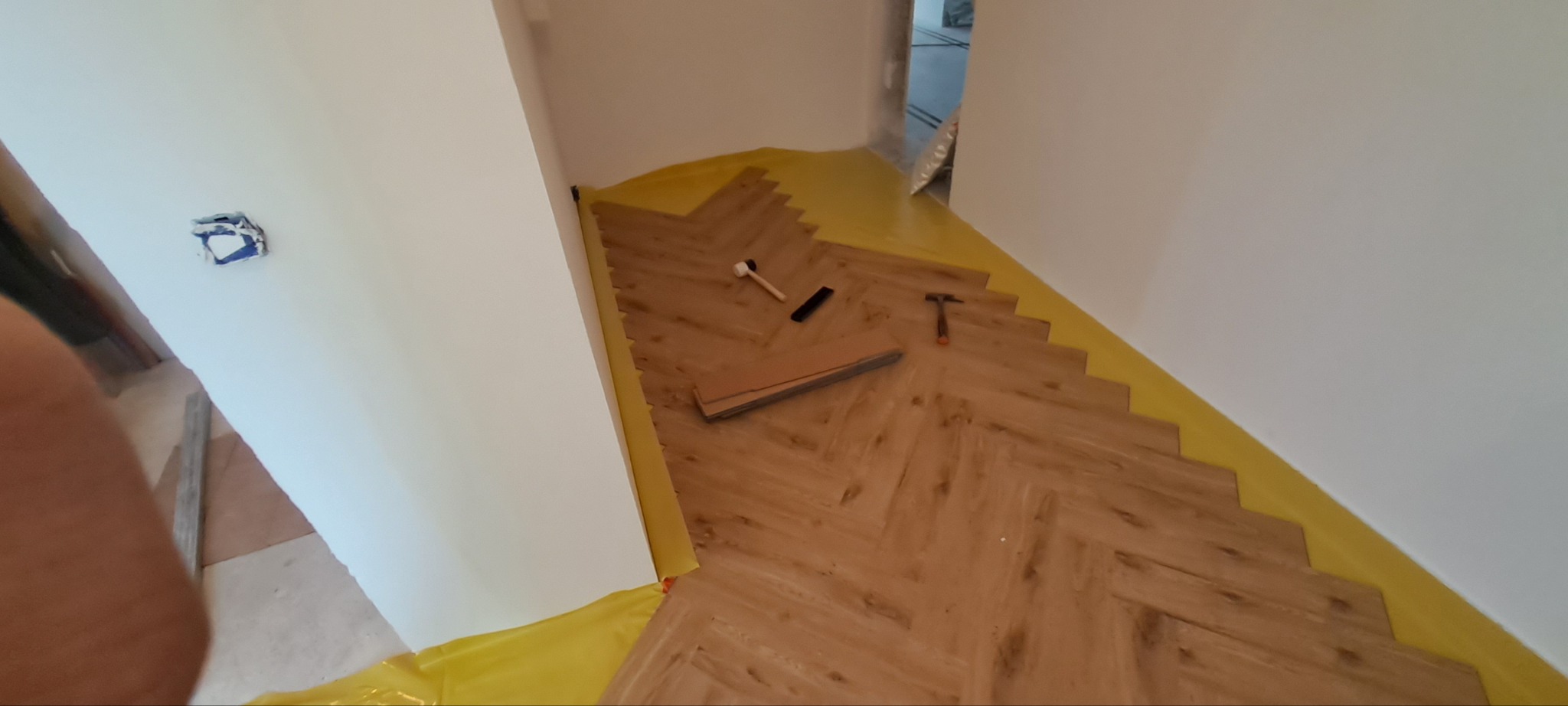 Układanie parkietu w jodełkę, widoczne narzędzia: młotek gumowy, dobijak, młotek stalowy. Podłoga zabezpieczona folią. Częściowo ułożony parkiet w jasnym odcieniu drewna.