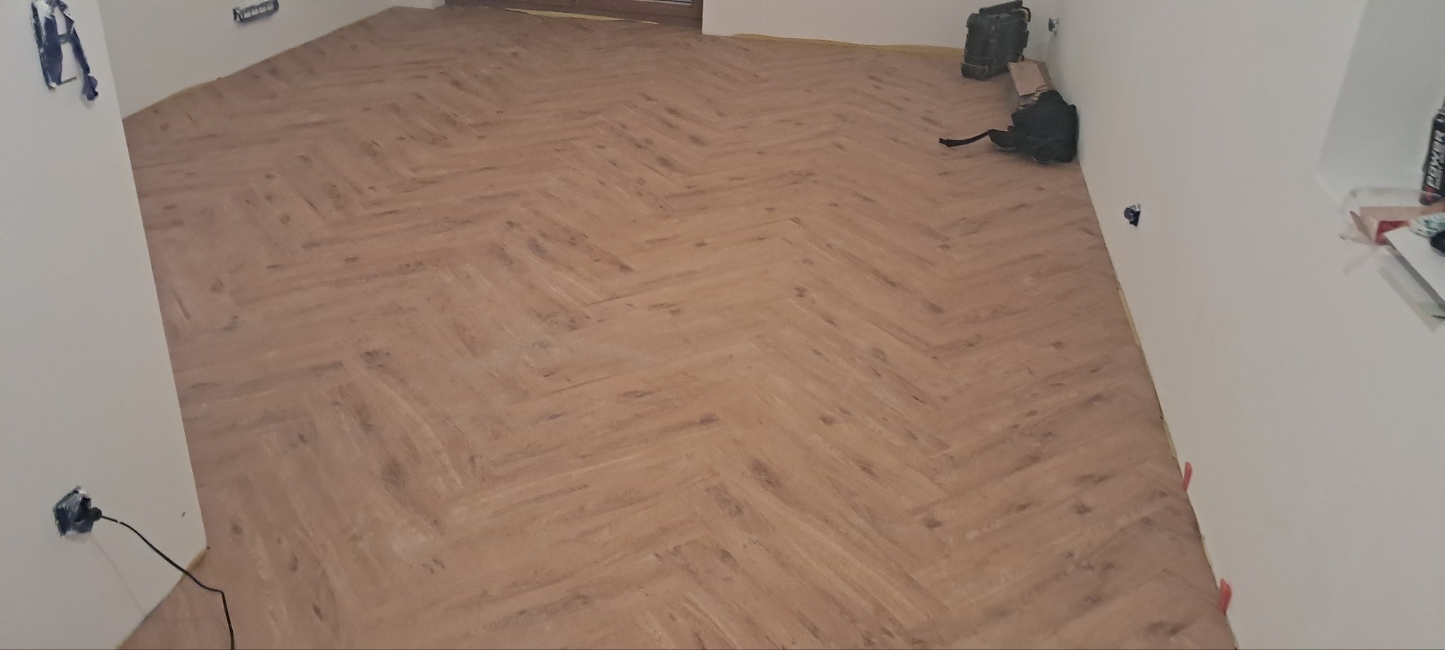 Ułożona podłoga z paneli w jodełkę, widok z góry. Widoczne narzędzia i akcesoria montażowe. Jasne ściany, gniazdko elektryczne.