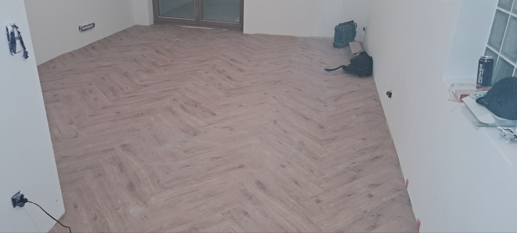 Ukończona podłoga z paneli ułożonych w jodełkę, widoczne gniazdka elektryczne i okno. Na parapecie puszka napoju energetycznego.