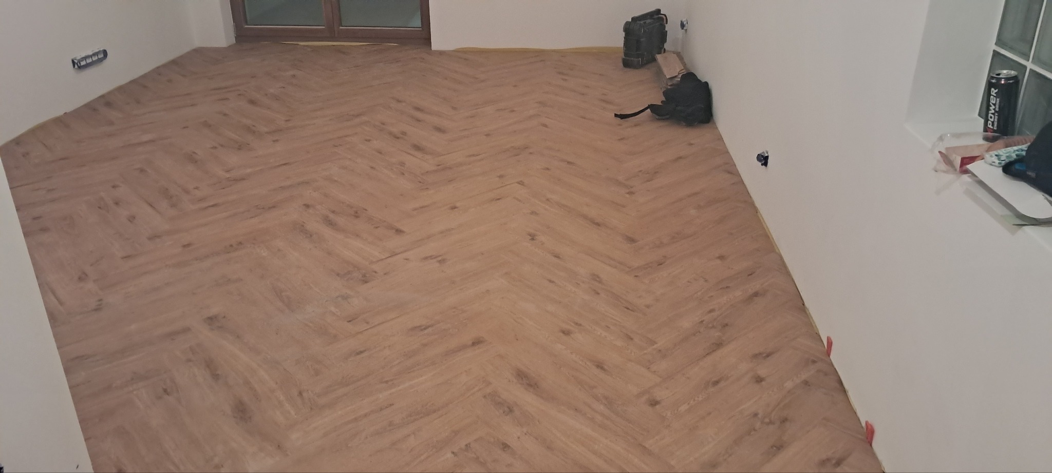 Ukończone układanie paneli podłogowych w jodełkę, widoczne narzędzia pracy przy ścianie, świeżo wykończone wnętrze.