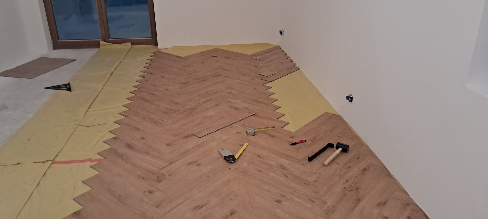 Układanie paneli w jodełkę na podkładzie. Widoczne narzędzia: miarka, młotek, kątownik. Częściowo ułożona podłoga w jasnym pomieszczeniu z oknem.