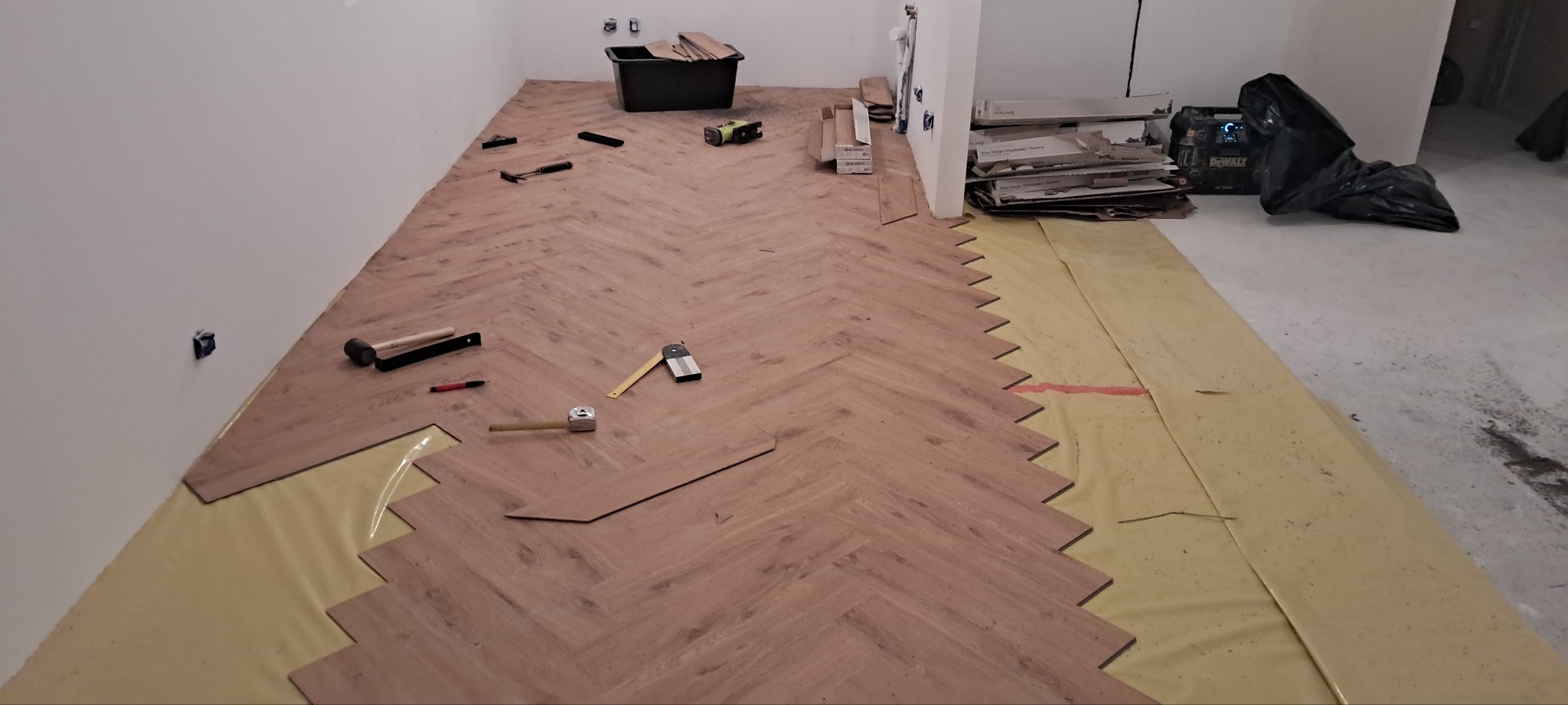Układanie parkietu w jodełkę francuską. Widoczne narzędzia, folia izolacyjna i częściowo ułożona podłoga. W tle kartony z panelami i czarny worek.