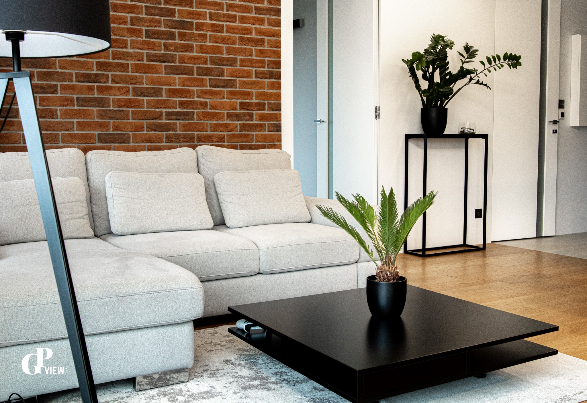 Jasnoszara sofa narożna z poduszkami na tle ściany z czerwonej cegły, czarny stolik kawowy z rośliną w doniczce, wysoka lampa podłogowa i drewniana podłoga tworzą nowoczesne wnętrze.