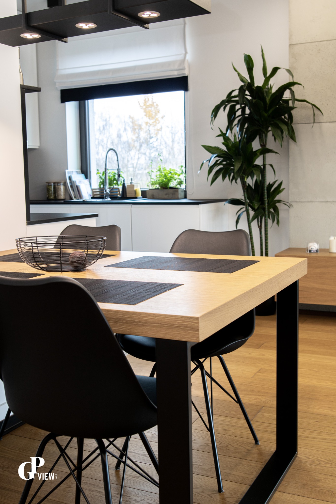 Jasny drewniany stół z czarnymi nogami i krzesłami w nowoczesnym wnętrzu, widok na okno z roślinami i minimalistyczną kuchnię.