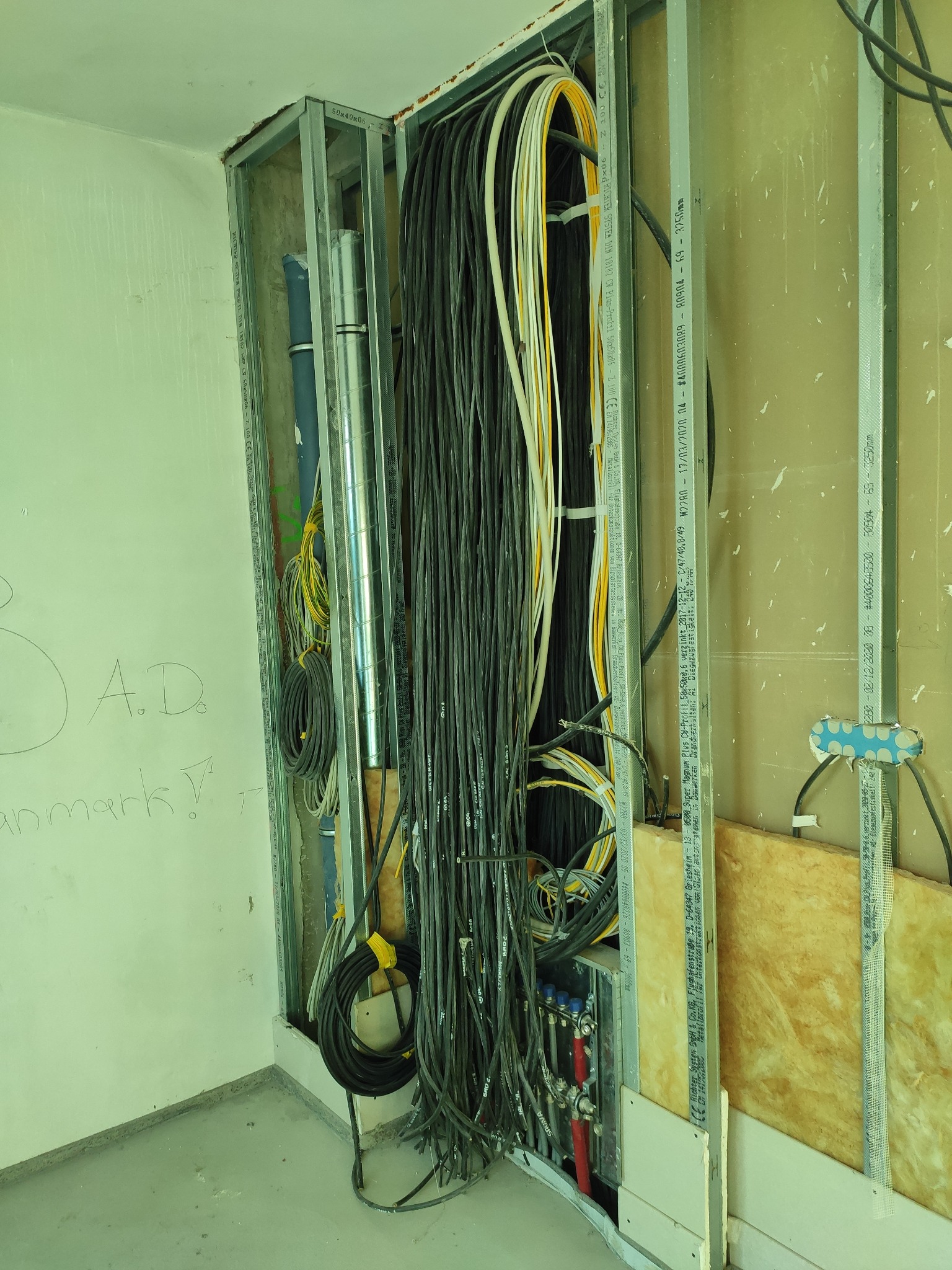 Szkielet metalowy pod zabudowę z widocznymi przewodami elektrycznymi i rurami instalacyjnymi w trakcie prac wykończeniowych, z fragmentem ściany z niedokończonymi napisami.