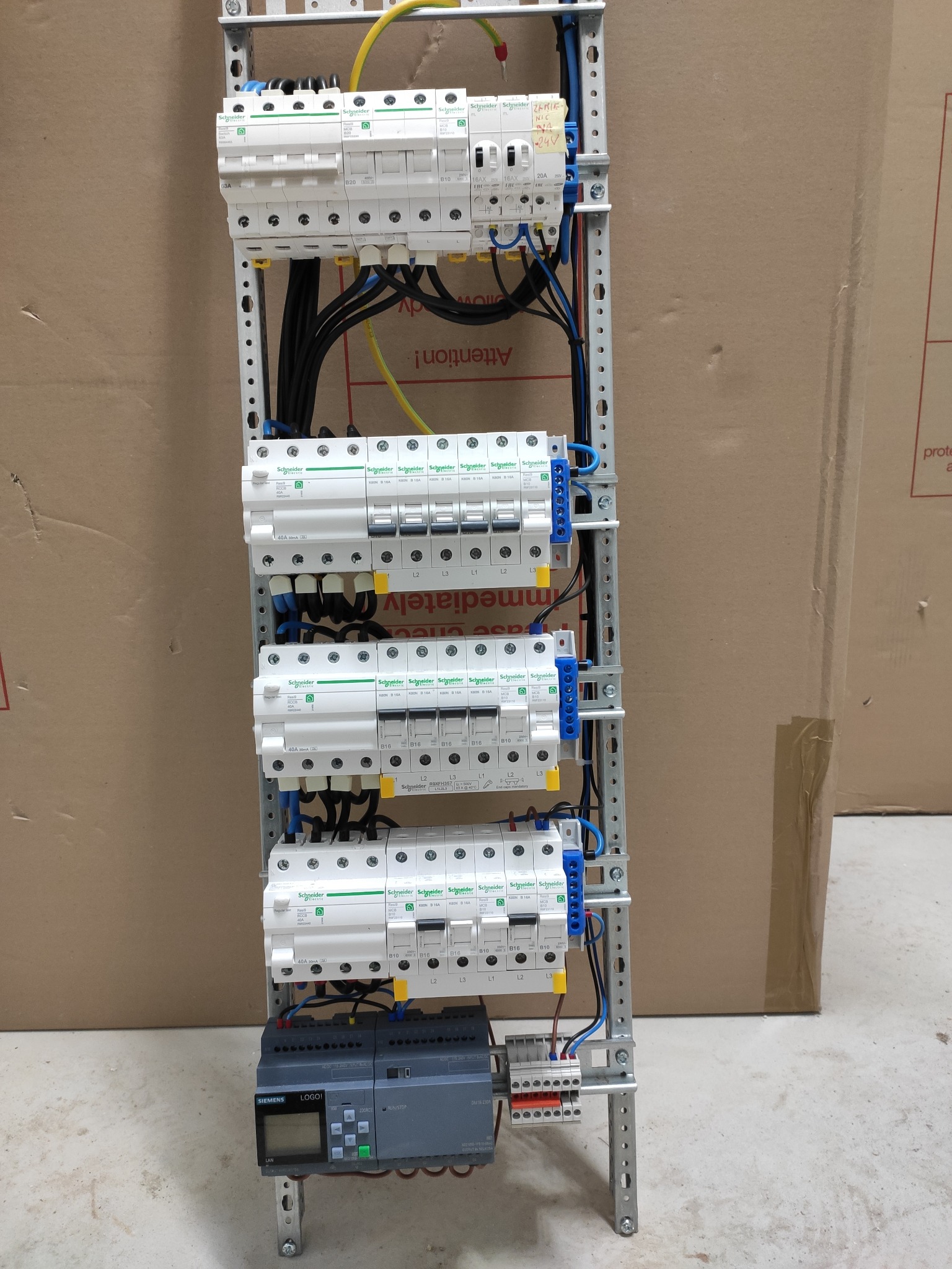 Rozdzielnica elektryczna z zamontowanymi bezpiecznikami i modułem sterującym Siemens LOGO!, widoczne okablowanie i oznaczenia, tło stanowi kartonowe opakowanie.
