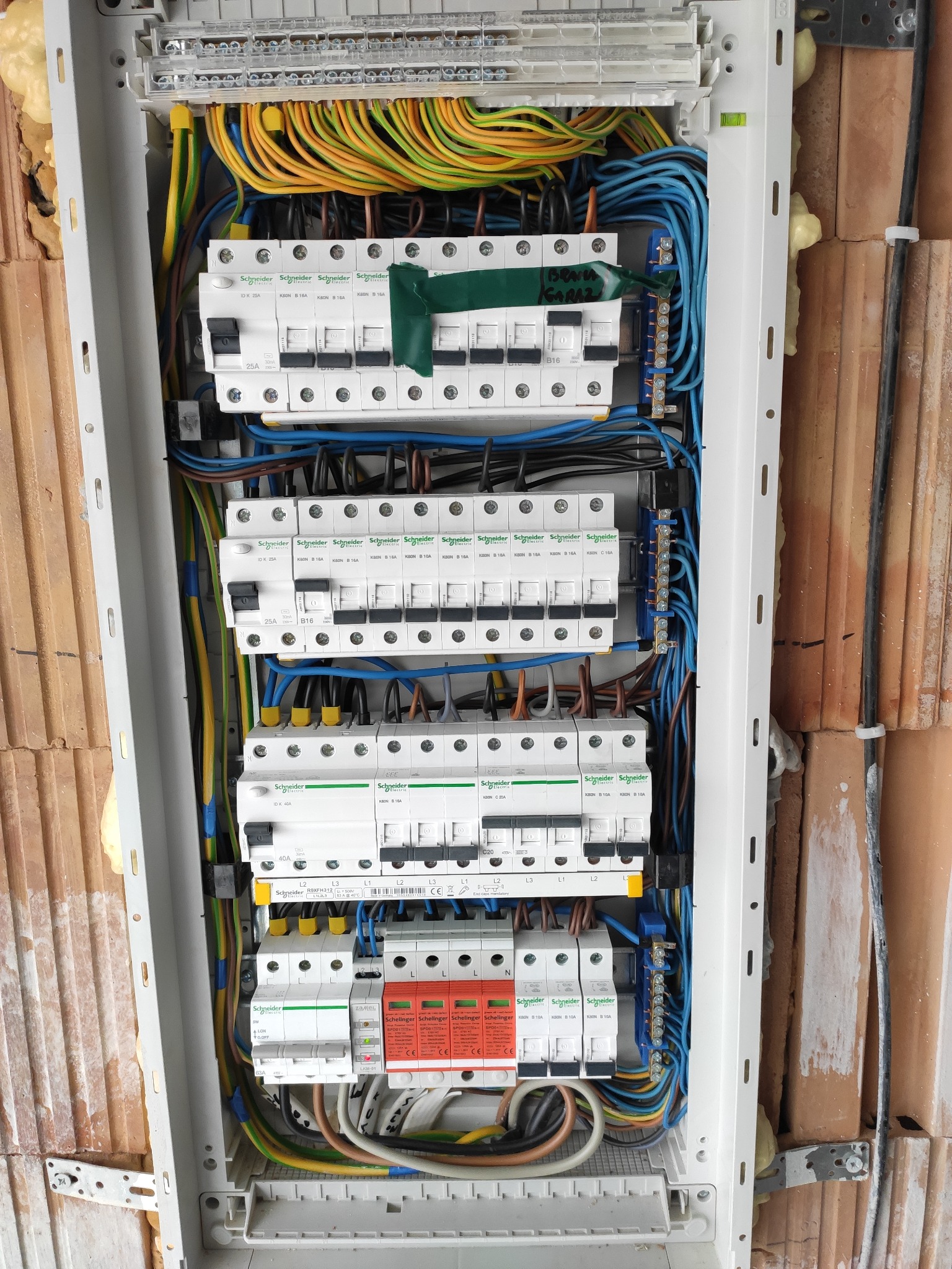 Otwarta skrzynka rozdzielcza z bezpiecznikami Schneider Electric, widoczne kolorowe przewody, instalacja w surowym stanie, ściana z cegły ceramicznej obok.