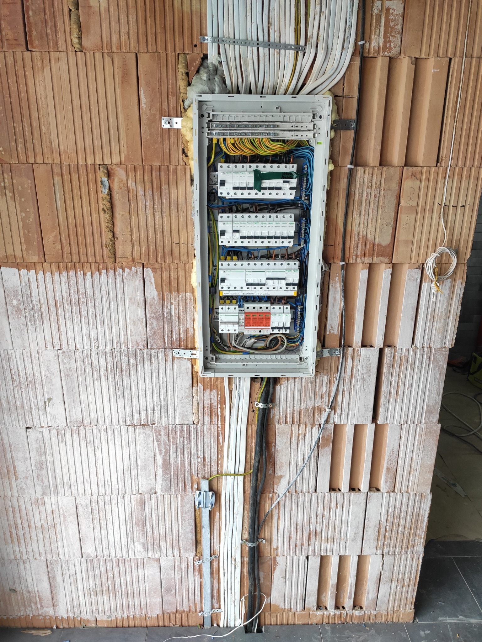 Rozdzielnica elektryczna zamontowana na ścianie z cegieł ceramicznych, widoczne kable i przewody w peszlach, instalacja w trakcie montażu.