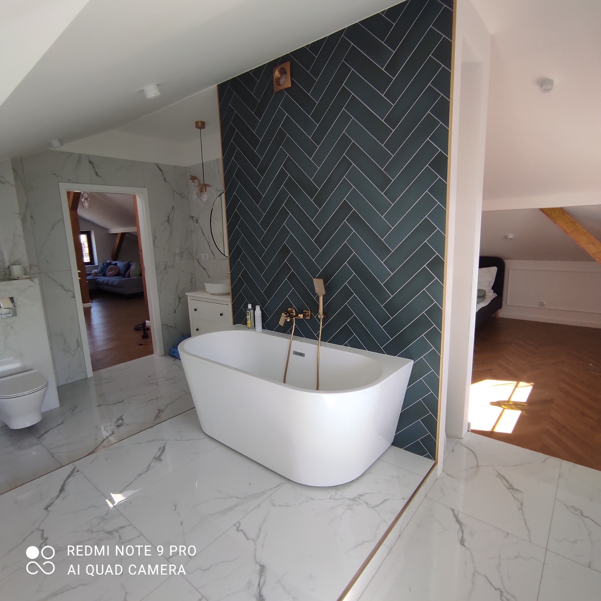 Nowoczesna łazienka w odcieniach bieli i szarości z akcentem ciemnozielonej ściany z płytek ułożonych w jodełkę, wolnostojąca wanna z złotą armaturą, marmurowe płytki podłogowe i widok na sąsiedni...