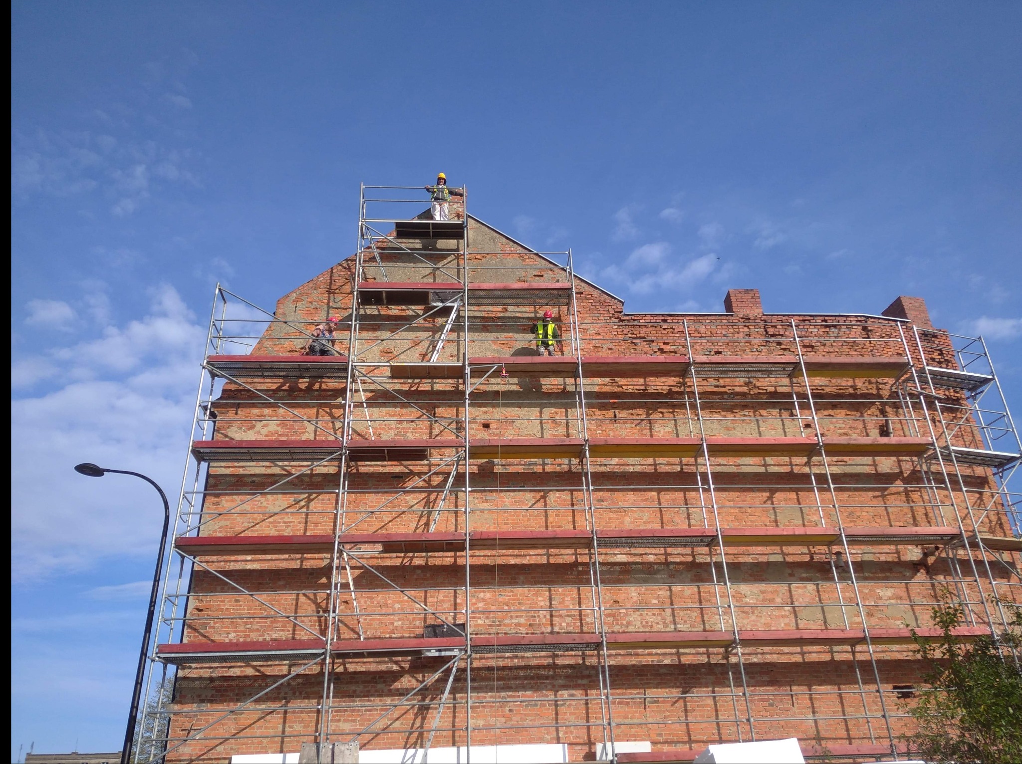 Wysoka ceglana kamienica w trakcie renowacji, otoczona rusztowaniami, z pracownikami w kaskach i kamizelkach odblaskowych na różnych poziomach, błękitne niebo w tle.
