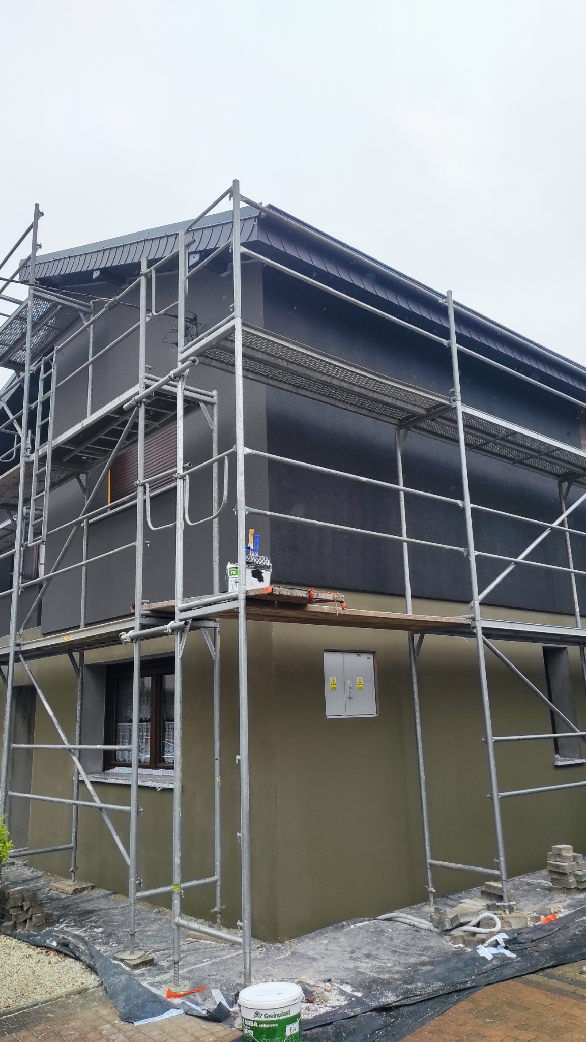 Dom jednorodzinny w trakcie prac elewacyjnych, widoczne rusztowania, wiadro z farbą Greinplast i pomalowana na szaro górna część budynku, dolna część w kolorze piaskowym, okno z firanką.