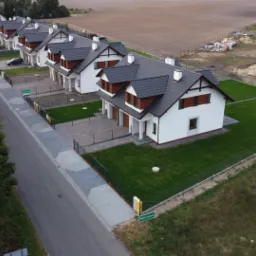 Widok z lotu ptaka na szereg nowo wybudowanych, identycznych domów jednorodzinnych z ciemnymi dachami i białymi elewacjami, z zagospodarowanymi trawnikami i chodnikami z kostki, obok widać plac...