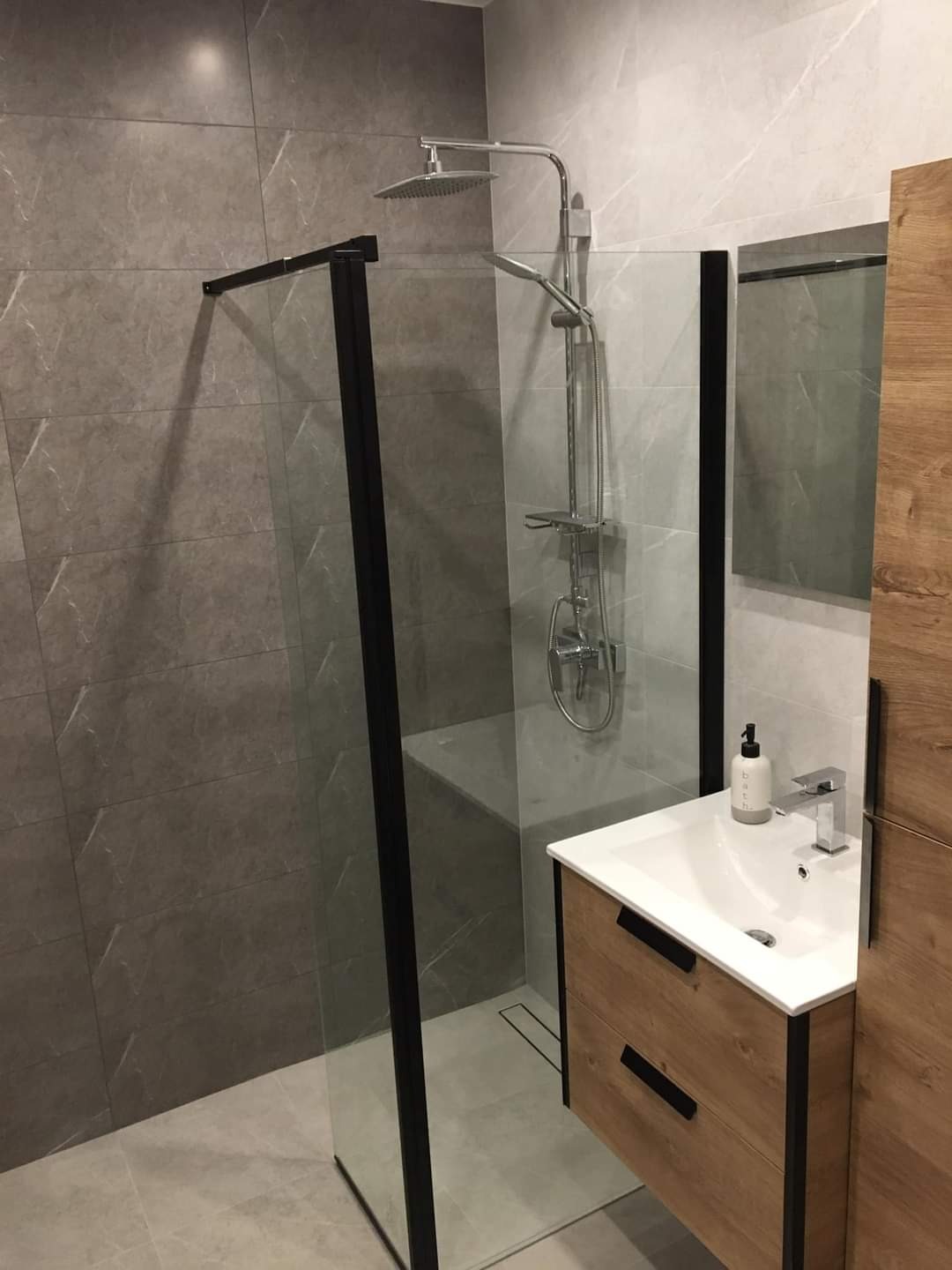 Nowoczesna łazienka z kabiną prysznicową typu walk-in z czarnymi ramami, umywalką nablatową na drewnianej szafce i szarą płytką imitującą marmur na ścianach.