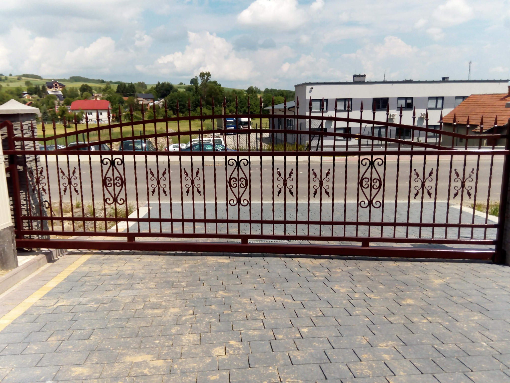 Przesuwna brama wjazdowa z metalowymi zdobieniami w kolorze ciemnej czerwieni, widok z poziomu kostki brukowej, w tle zabudowa wiejska i błękitne niebo.