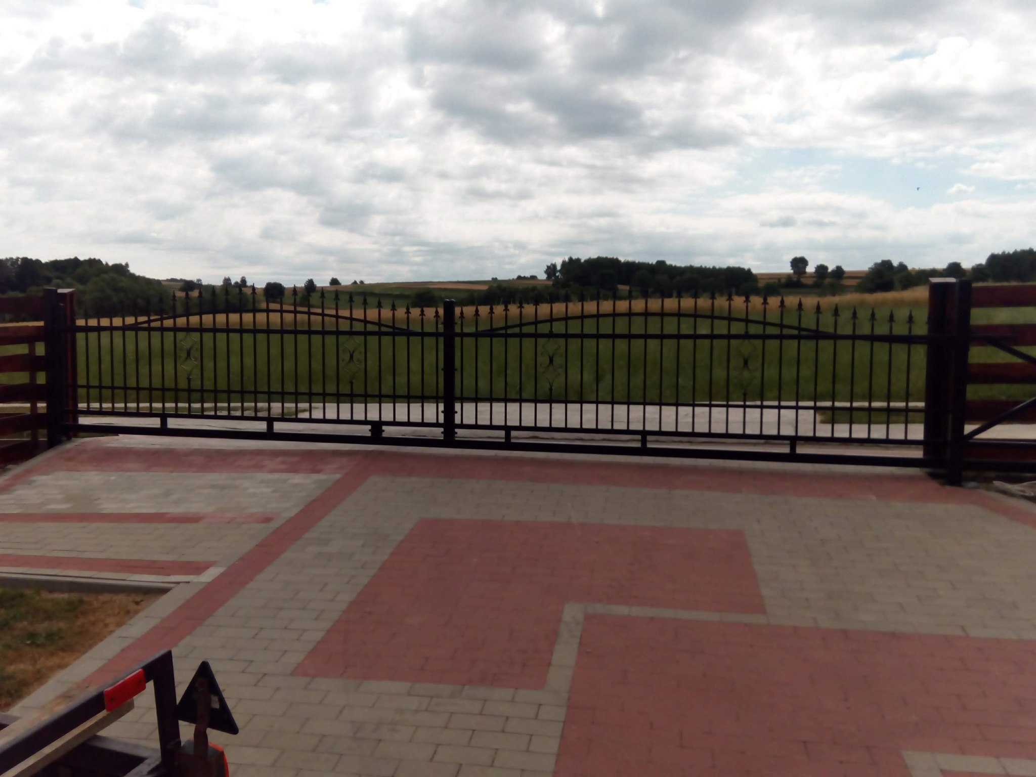 Czarna, metalowa brama wjazdowa z drewnianymi słupkami na posesji z kostki brukowej, w tle widok na pola i las pod pochmurnym niebem.