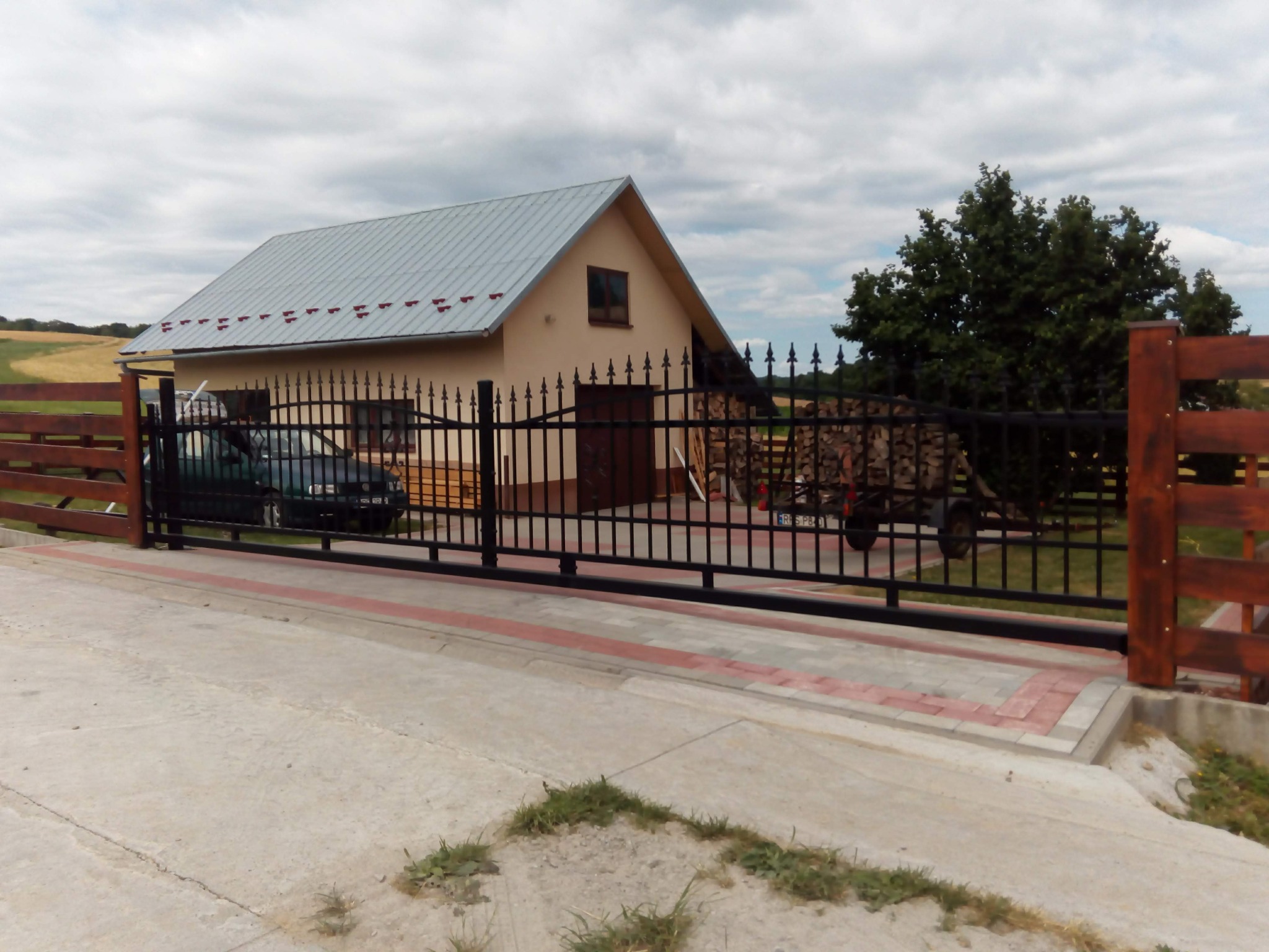 Czarna, przesuwna brama wjazdowa z metalowymi prętami na posesji z domem, samochodem i stertą drewna na przyczepce w tle.