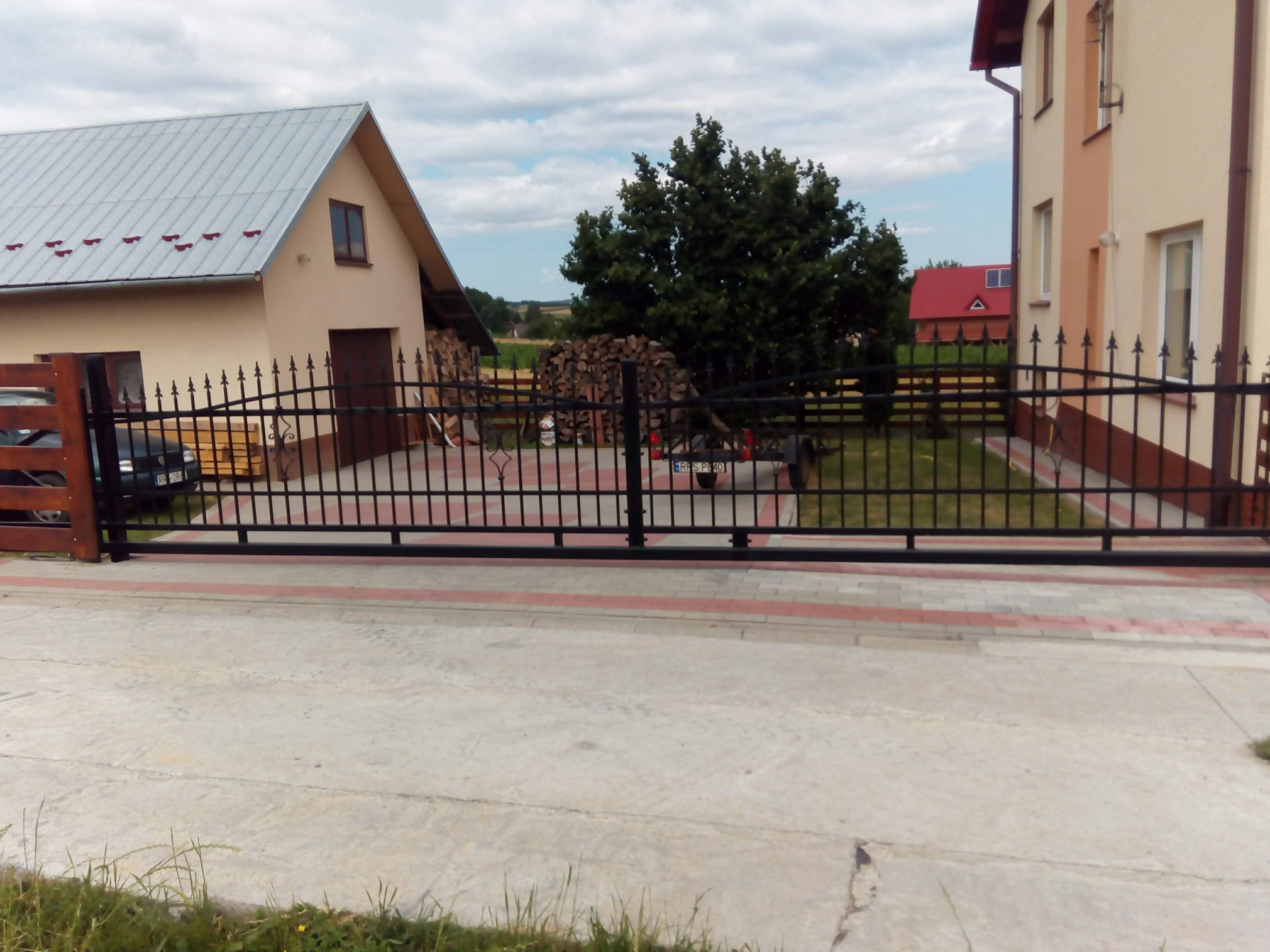 Czarna, przesuwna brama wjazdowa z metalowymi prętami na posesji z dwoma budynkami mieszkalnymi i stosem drewna w tle.