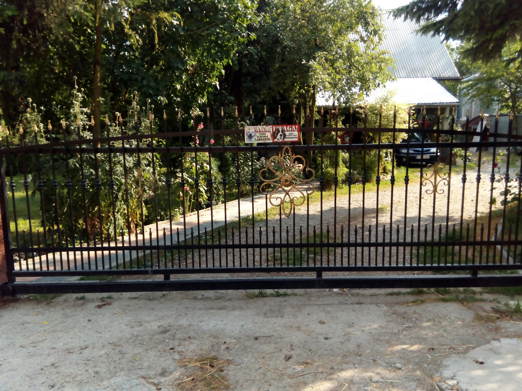 Czarna, przesuwna brama wjazdowa z metalowymi prętami i ozdobnym, złotym elementem pośrodku, widoczna z zewnątrz posesji w Łączkach Kucharskich, z tabliczką informującą o ochronie posesji przez psy.