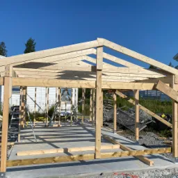 Konstrukcja szkieletowa z drewna, prawdopodobnie altany lub garażu, na betonowej wylewce, w trakcie budowy, widoczne elementy mocujące i narzędzia w tle.