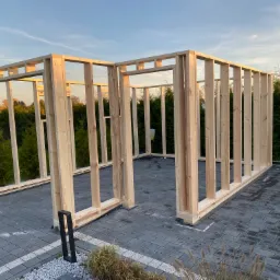 Konstrukcja szkieletowa budynku w trakcie budowy, widoczne drewniane ramy na szarej kostce brukowej, w tle zieleń i błękitne niebo o zachodzie słońca.