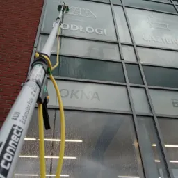 Czyszczenie szklanej fasady budynku z napisem 'PODŁOGI', 'ŁAZIENKI', 'OKNA' za pomocą teleskopowej lancy zakończonej szczotką, widoczne smugi wody.