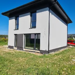 Dwukondygnacyjny dom z jasną elewacją i ciemną stolarką okienną, widoczne pęknięcia na tynku elewacyjnym, trawa na pierwszym planie, błękitne niebo w tle.