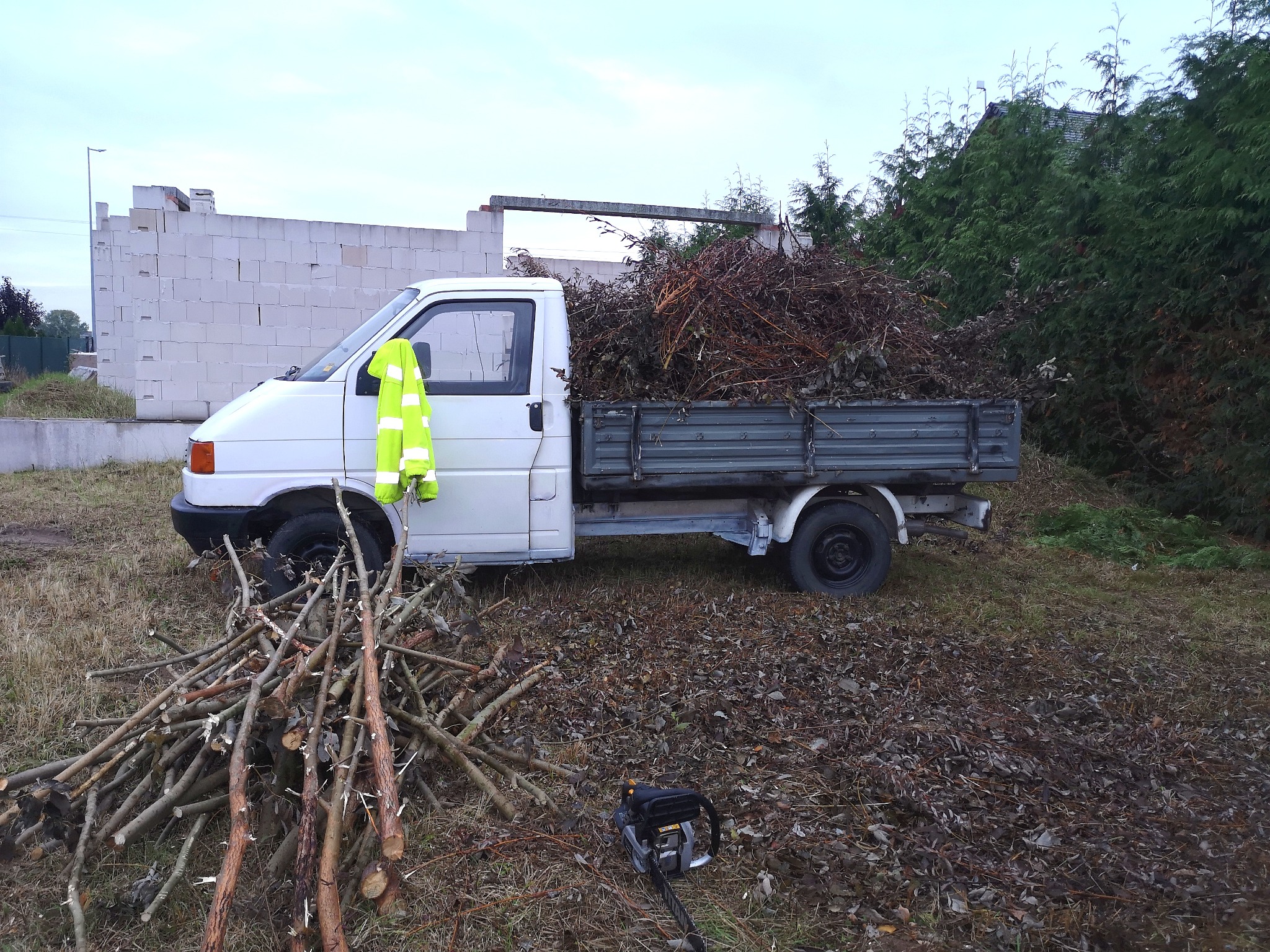 Biały samochód dostawczy z przyczepką wypełnioną gałęziami i liśćmi, żółta kamizelka odblaskowa na drzwiach, obok leżą pocięte gałęzie i piła łańcuchowa, w tle mur z pustaków.