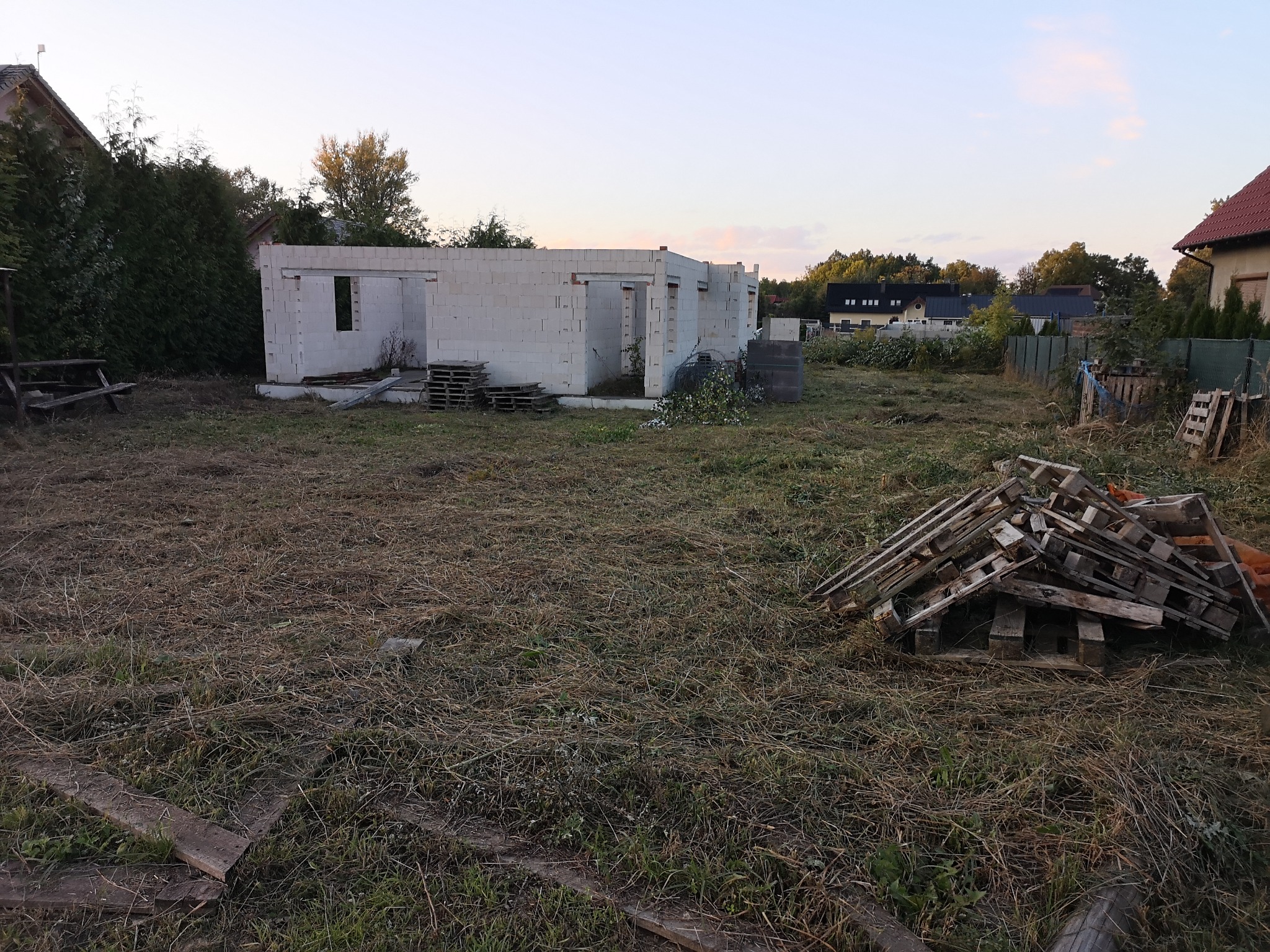 Stan surowy otwarty domu jednorodzinnego w Czarnkowie, widoczne ściany z bloczków i stosy drewnianych palet na zarośniętej trawie, w tle zabudowa mieszkalna.