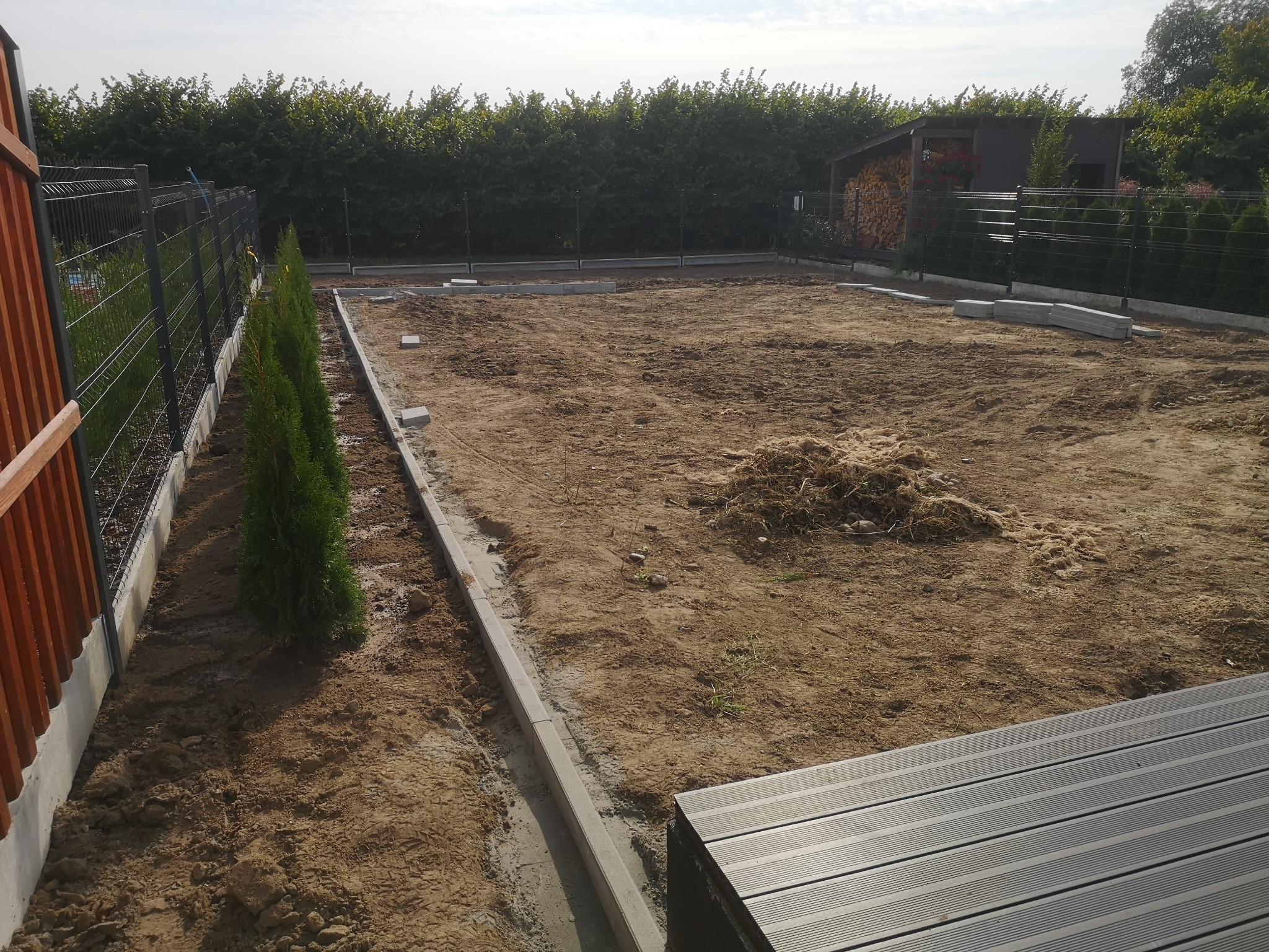 Przygotowany teren pod przyszły ogród: wyrównana ziemia, obrzeża z kostki, posadzone małe tuje wzdłuż ogrodzenia, w tle drewniana wiata na drewno.