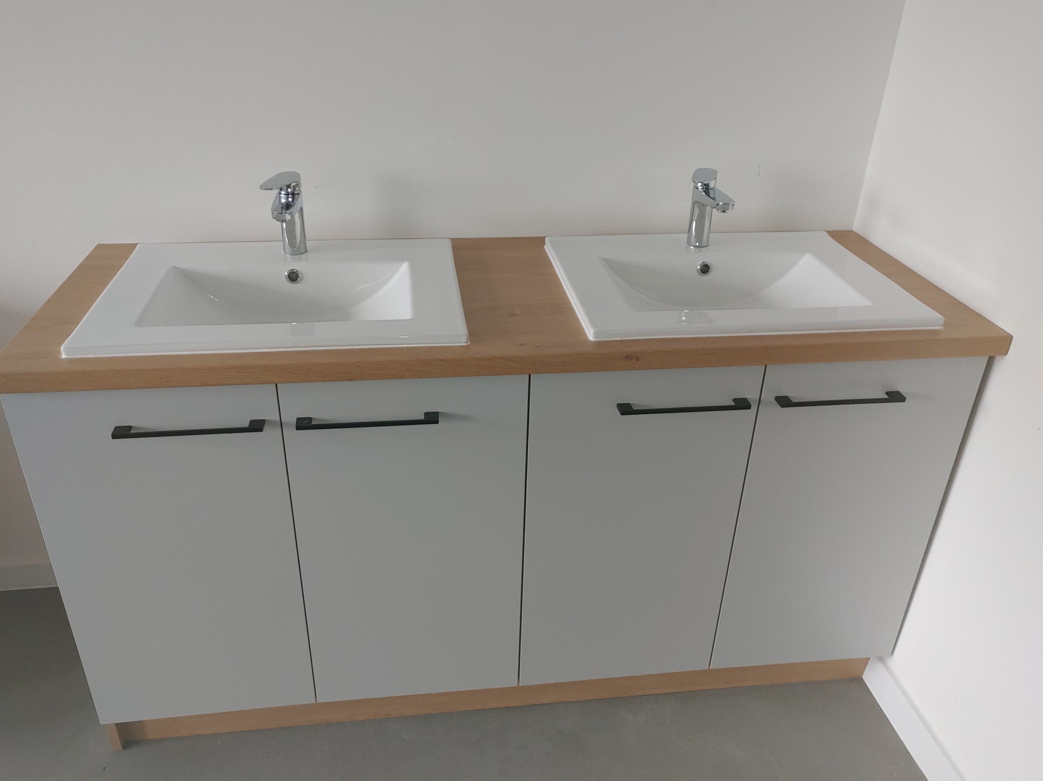 Dwie białe umywalki wpuszczone w drewniany blat, zamontowane na szarej szafce z czarnymi uchwytami, minimalistyczny design.