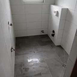 Wnętrze łazienki w trakcie remontu: ułożone szare płytki podłogowe, białe płytki ścienne, widoczne przyłącza wodne i otwór na odpływ toalety podtynkowej, okno w górnej części ściany.