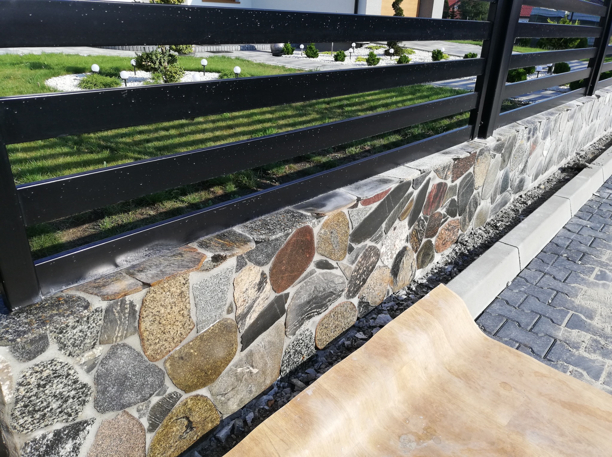 Kamienny murek z czarnym metalowym ogrodzeniem na tle zielonego trawnika z oświetleniem ogrodowym, widoczna krawędź chodnika i fragment deski.