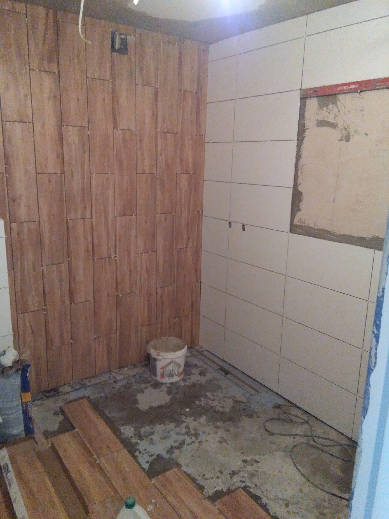 Łazienka w trakcie remontu: ściana wyłożona płytkami imitującymi drewno, sąsiadująca ze ścianą z jasnymi, prostokątnymi płytkami; na podłodze widoczne płytki do montażu oraz wiadro, wskazujące...