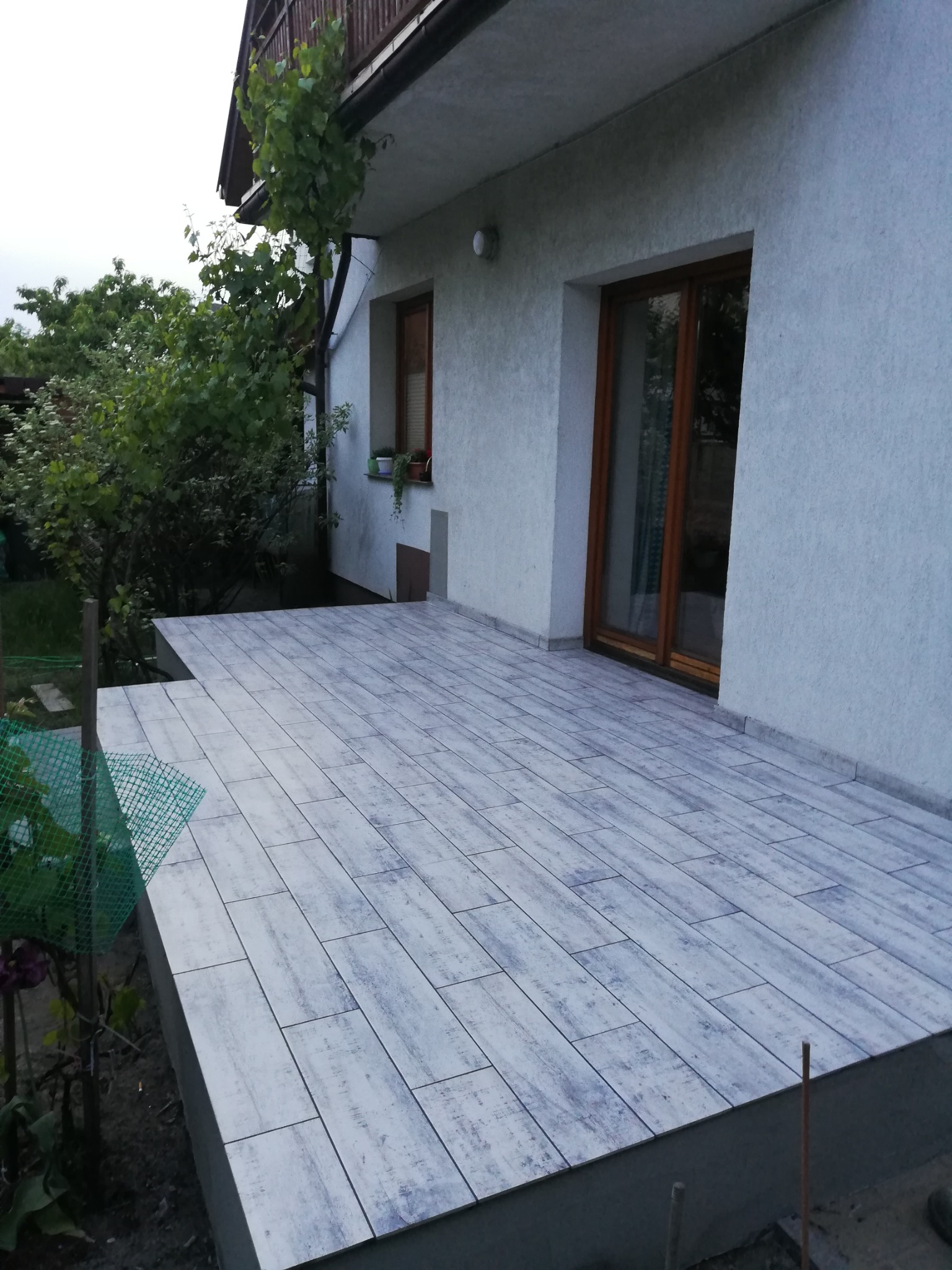 Wykończony taras przed domem z płytkami imitującymi drewno, widok na elewację z oknami i pnączami.
