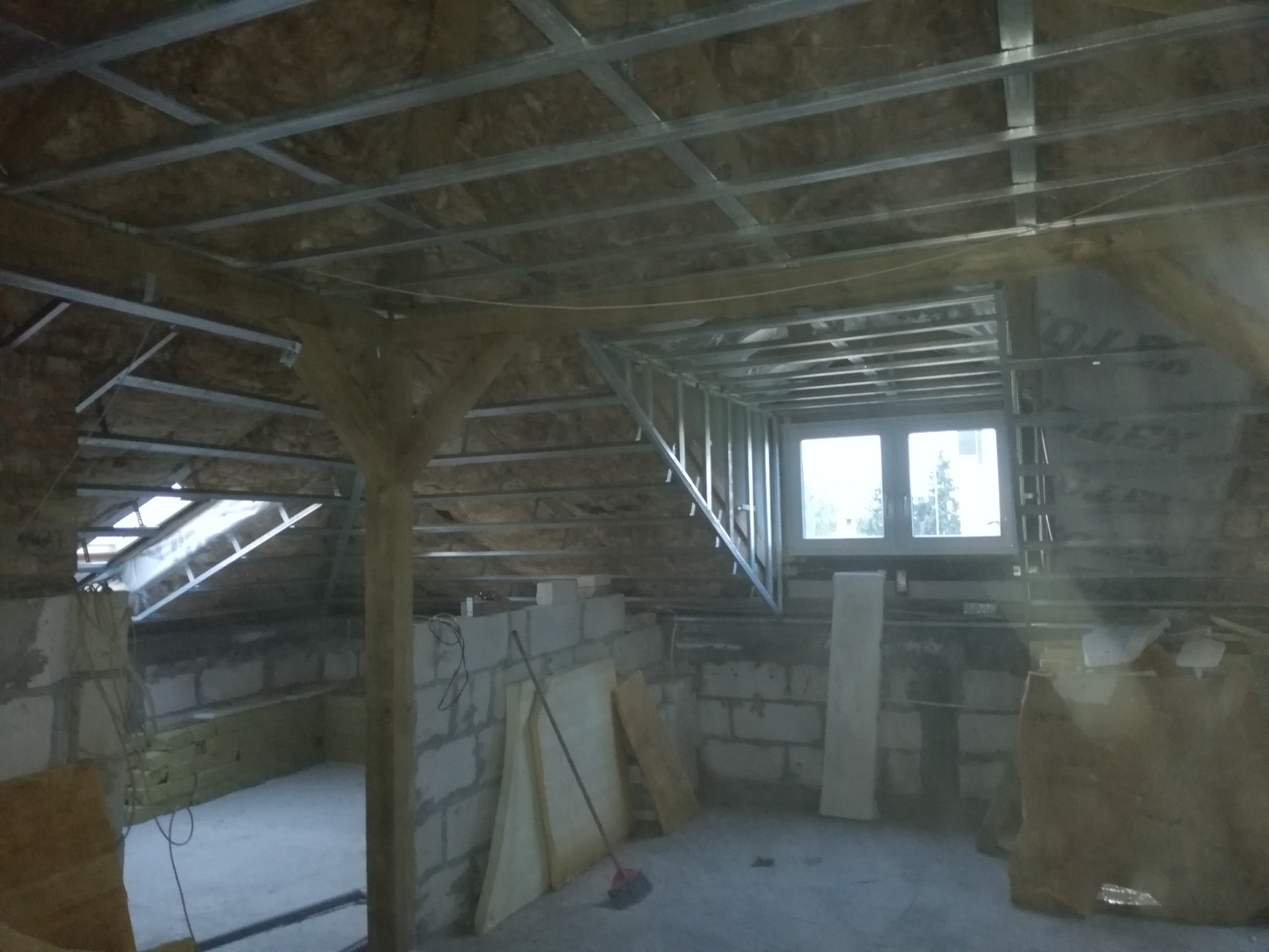 Poddasze w trakcie prac wykończeniowych: widoczne metalowe profile sufitowe, ocieplenie, ściany z bloczków, drewniany słup, okno i materiały budowlane.