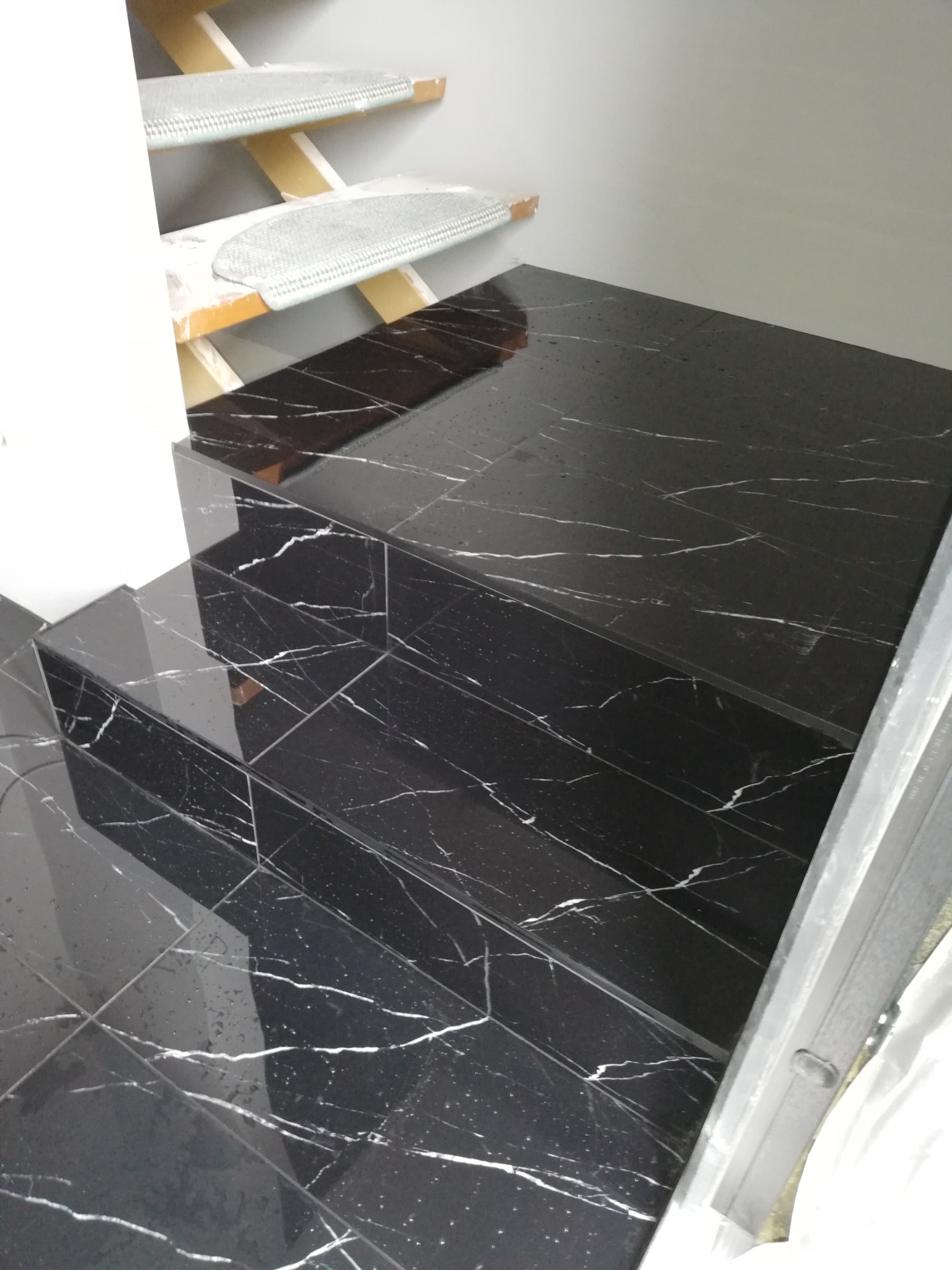 Schody wyłożone błyszczącymi, czarnymi płytkami imitującymi marmur z białymi żyłami, widok z góry, częściowo widoczne drewniane stopnie schodów z szarymi nakładkami antypoślizgowymi.