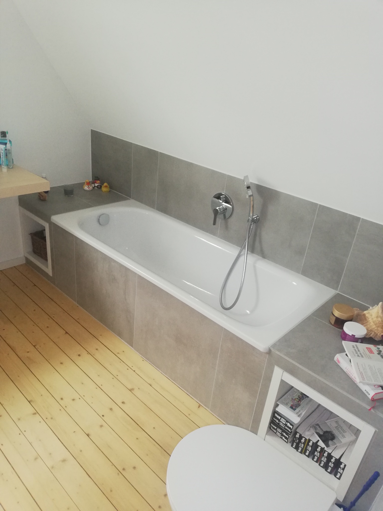 Wykończona łazienka na poddaszu z drewnianą podłogą, zabudowaną wanną w szarych płytkach, baterią wannową na ścianie i półkami na książki po bokach wanny.