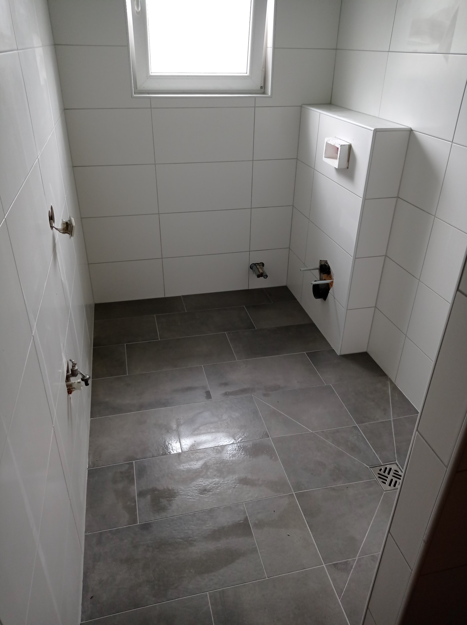 Wnętrze łazienki w trakcie remontu: ułożone szare płytki podłogowe, białe płytki ścienne, widoczne przyłącza wodne i otwór na odpływ toalety podtynkowej, okno w górnej części ściany.